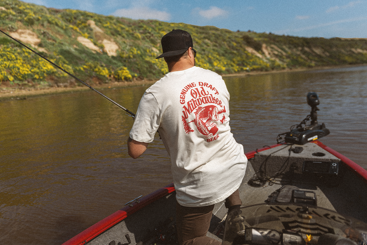 Man fishing from a boat on a river with a scenic background wearing an Old Milwaukee shirt.