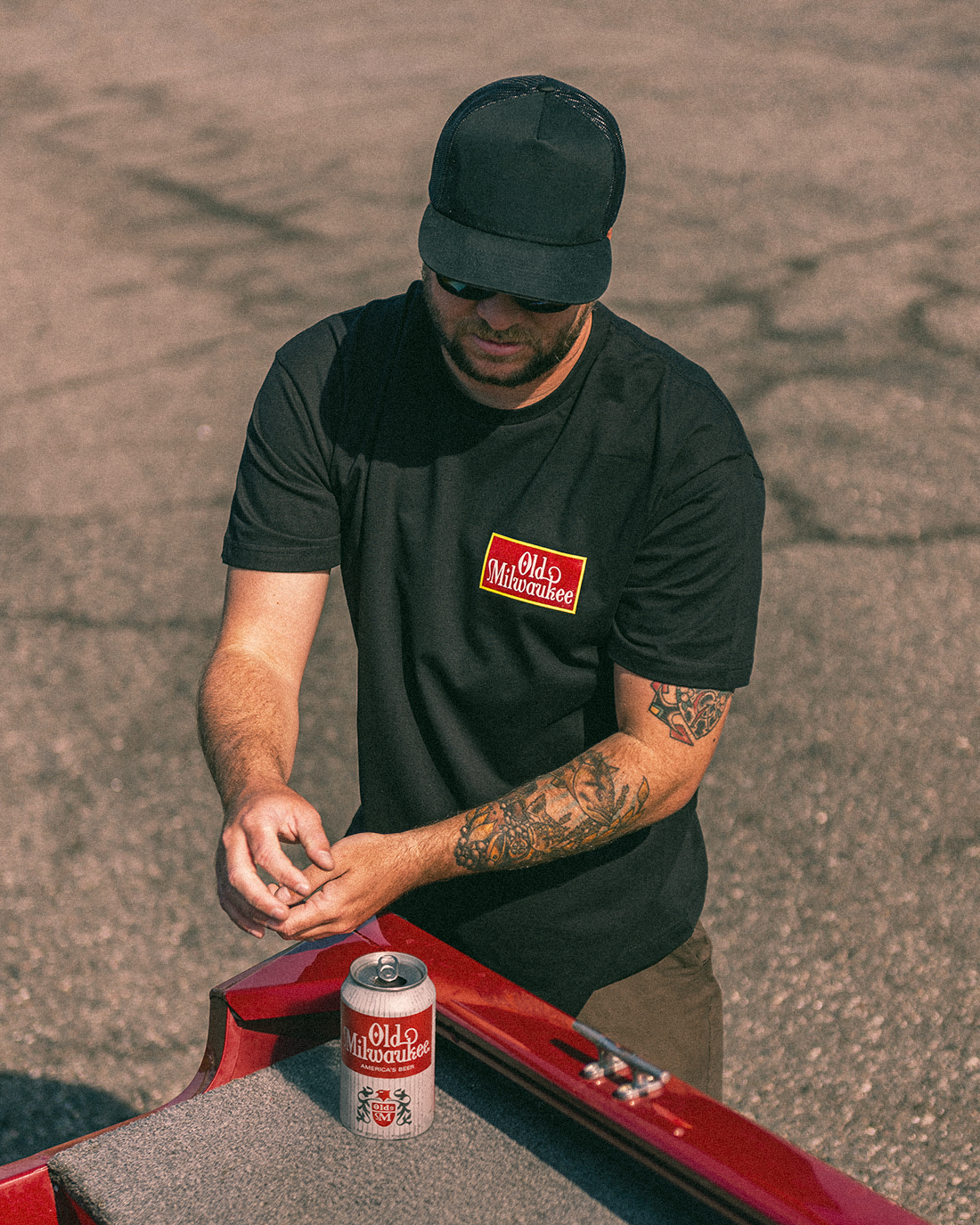Man in black t-shirt and cap with tattooed arm, standing next to a red car with Old Milwaukee beer can.