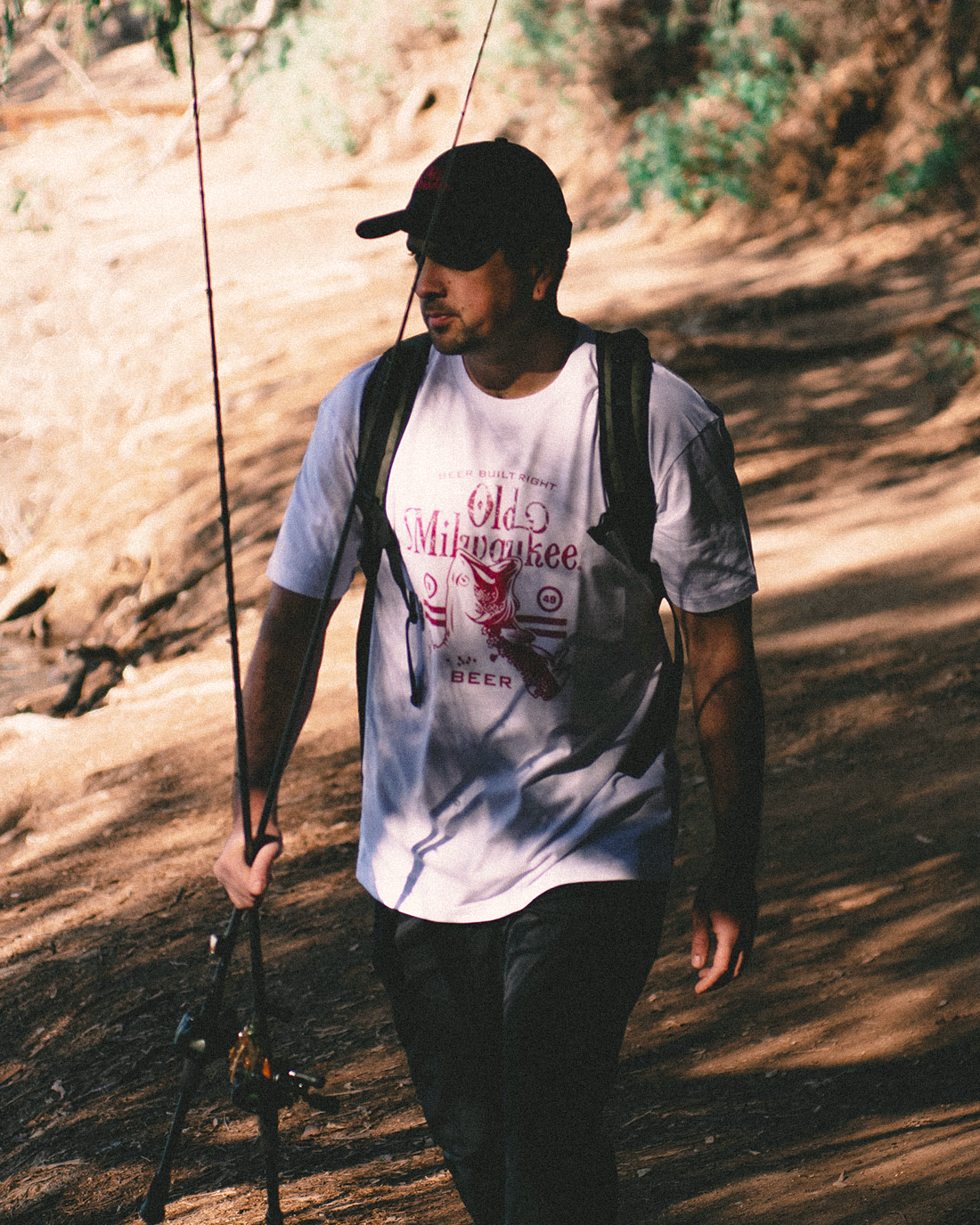 Man walking outdoors with a fishing pole, wearing a white t-shirt and black cap.